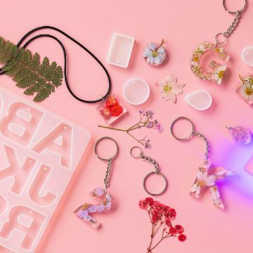Show details for Alphabet Mold A collection of floral resin keychains and necklaces on a pink surface. Various dried flowers are embedded in letters and numbers. Nearby are a silicone mold and loose dried flowers, adding to the craft aesthetic.