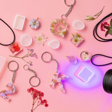 Show details for Keychain Findings Kit Various resin crafts are scattered on a pink surface, including keychains and necklaces with embedded flowers and letters. A UV light and dried flowers are also visible, suggesting a crafting setup.