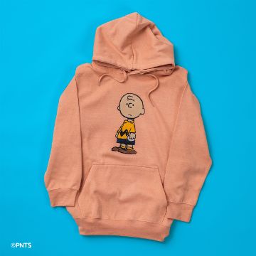Show details for Peanuts - Charlie Brown A peach-colored hoodie features a printed cartoon character wearing a yellow shirt with a zigzag pattern. The hoodie is laid flat on a bright blue background.
