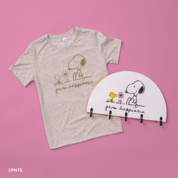 Show details for Peanuts - Grow Happiness A beige T-shirt featuring a cartoon dog and bird with the text "grow happiness" is laid on a pink background. Next to it is a matching semicircular wall hook with the same design.