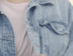 A person wearing a light pink top and a light blue denim jacket, with a chain necklace visible around their neck. The photo is cropped to show only the upper chest and shoulders.