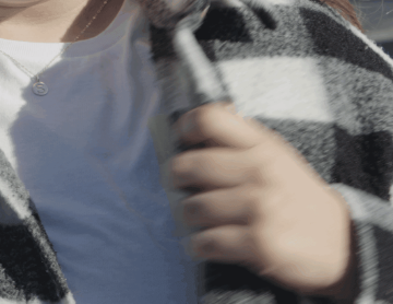 Show details for Sun Ink Single A person wearing a white shirt and black-and-white plaid scarf holds the scarf closed. They have a necklace with a round pendant featuring the letter “S.” The image is closely cropped to show the upper chest and hand.