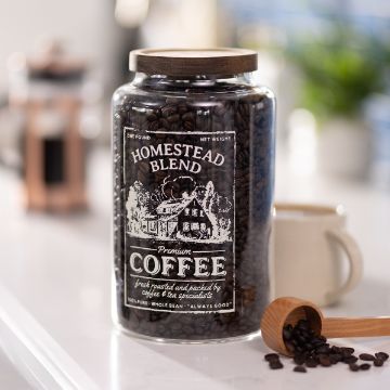 Show details for Homestead Blend A large glass jar labeled "Homestead Blend Premium Coffee" filled with coffee beans sits on a countertop, with a wooden scoop holding beans beside it and a blurred coffee mug in the background.