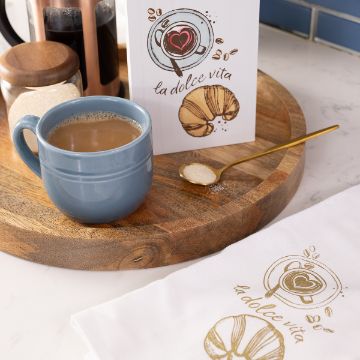Show details for La Dolce Vita A blue mug of coffee sits on a wooden tray beside a gold spoon with sugar, a jar, a French press, and a card featuring coffee and pastry illustrations with the words “la dolce vita.” A matching napkin is nearby.