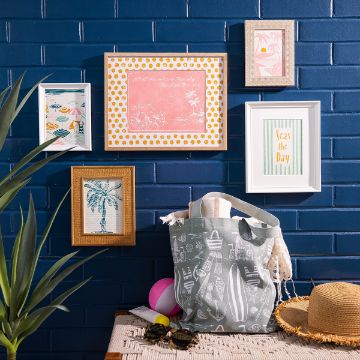 Show details for Ocean View A gray beach bag with towels and a hat sits on a table in front of a dark blue brick wall decorated with five framed, colorful summer-themed prints. A green plant and a straw hat are also visible.