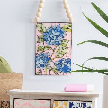 Show details for Stained Glass Hydrangea A decorative hanging art piece with blue hydrangea flowers and green leaves on a pink background, displayed on a wall above a shelf with plants and small decor items.