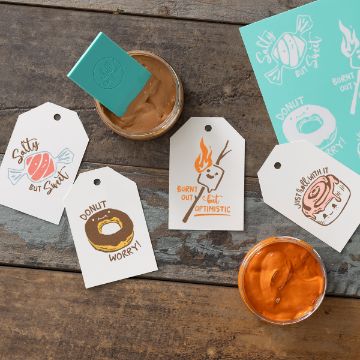 Show details for Donut Worry Gift tags with playful dessert illustrations and puns sit on a rustic wooden table beside containers of brown and orange spreads, a teal envelope, and a sheet of matching stickers.