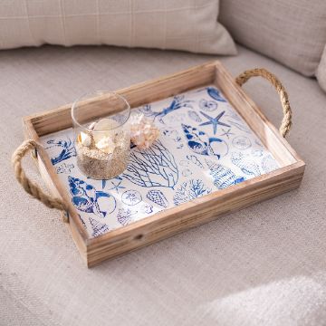 Show details for By The Seashore A wooden tray with rope handles, lined with a blue and white seashell and coral design. On the tray are a glass filled with sand and seashells and a couple of loose seashells, resting on a beige couch.