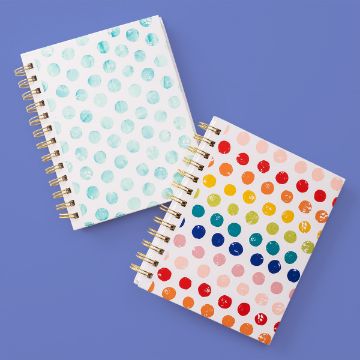 Show details for Watercolor Dot Pattern Two spiral-bound notebooks with polka dot covers lie on a purple surface. One notebook has blue dots on a white background; the other has multicolored dots on a light pink background.