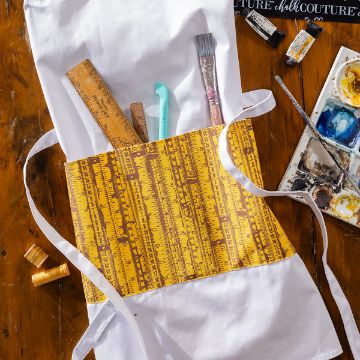Show details for Vintage Ruler Pattern A white apron with a yellow pocket featuring ruler designs holds paintbrushes and rulers. It is laid on a wooden table with sewing thread, paint tubes, and a watercolor palette nearby.
