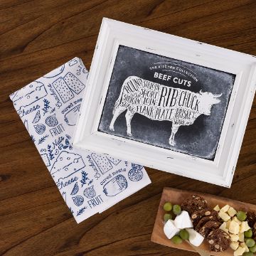 Show details for Beef Cuts A framed beef cuts diagram sits on a wooden table next to a folded, food-themed cloth. Nearby, a wooden board holds assorted cheeses, grapes, crackers, and nuts.