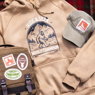 Show details for Hide & Seek Champion Tan hoodie with a "Hide & Seek World Champion" Sasquatch graphic, a khaki cap with a patch, and a brown backpack with patches, all arranged on a red and brown plaid blanket.