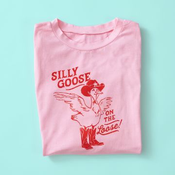 Show details for Silly Goose A folded pink T-shirt with a red illustration of a goose wearing a cowboy hat and boots, and the words "SILLY GOOSE ON THE Loose!" printed above and beside the goose. The shirt is on a light blue background.