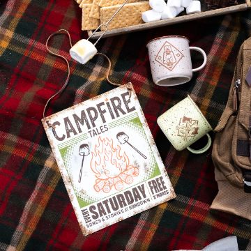 Show details for Campfire Tales A metal "Campfire Tales" sign with marshmallow graphics lies on a plaid blanket, beside two mugs, a marshmallow on a stick, a tray of graham crackers and marshmallows, and a tan backpack.