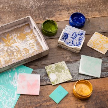 Show details for Floral Melody Assorted decorative ceramic tiles with botanical prints are arranged on a rustic wooden surface beside open jars of paint, a wooden tray with gold designs, and small green and blue cards.