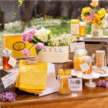 Show details for Busy Bees A bee-themed outdoor table display with honey jars, fresh flowers, honey dippers, yellow accents, and bee decorations. The setup includes labeled jars, honeycomb-patterned towels, and a “BUZZ” flower arrangement.