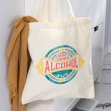 Show details for Everyday Tote Bag A cream-colored tote bag with a colorful vintage-style label that reads "May contain alcohol" hangs on a white cabinet, with a tan scarf draped over the handle.