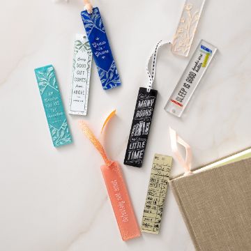 Show details for Bookmark Mold (1 1/8" x 5") A collection of nine colorful bookmarks with various designs and inspirational quotes, some with ribbons, are spread out next to an open beige book on a light surface.