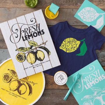 Show details for Slightly Bitter A flat lay displays lemon-themed items: two "Fresh Lemons" signs, a navy baby onesie with "Slightly Bitter" and a green lemon, matching paper cards, a jar of green paste, and a container of black ink.