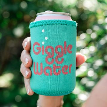 Show details for Giggle Water A hand holding a can wrapped in a teal drink sleeve with pink text that reads "Giggle Water," against a blurred, green outdoor background.