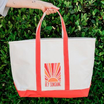Show details for Hey Sunshine A hand holds a cream tote bag with red handles and trim. The bag features a retro sunrise graphic and the text “HEY SUNSHINE.” Green leafy bushes are in the background.