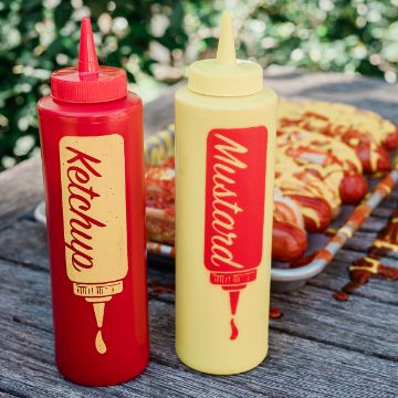 Show details for Squeeze Me Two squeeze bottles labeled ketchup and mustard sit on a wooden table outdoors, with a tray of hot dogs topped with ketchup and mustard in the blurred background.