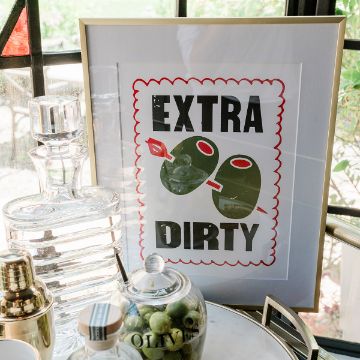 Show details for Extra Dirty A framed print with two olives and a toothpick reads "EXTRA DIRTY." It sits on a bar cart beside a decanter, cocktail shaker, glassware, and a jar labeled "OLIVES." Sunlight streams through nearby windows.