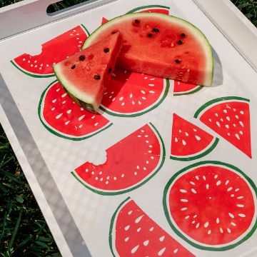 Show details for One In A Melon A white tray with watermelon slice illustrations holds two real watermelon wedges, one placed over the drawings. The tray rests on green grass, and sunlight creates a patterned shadow across the scene.