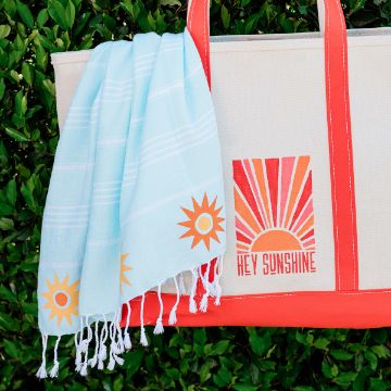 Show details for Solar-ious A canvas tote bag with orange handles and a "Hey Sunshine" sunburst design stands against green leaves. A light blue towel with orange sun motifs is draped over the bag.