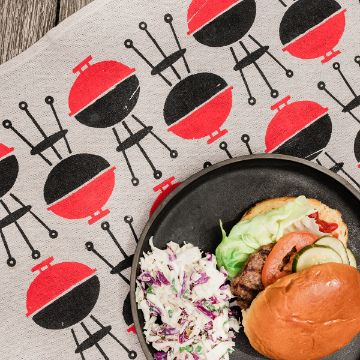 Show details for Hot Off The Grill A plate with a hamburger and coleslaw sits on a cloth decorated with a repeating pattern of red and black barbecue grills and chairs.