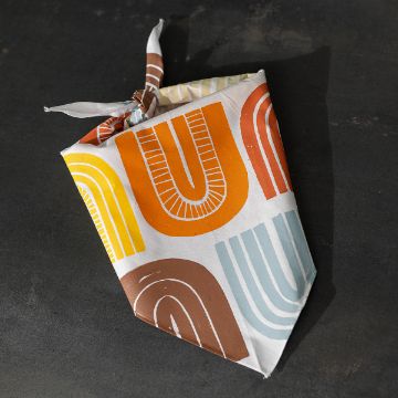 Show details for Check And Mate A folded cloth napkin with a knot, featuring a colorful geometric pattern in yellow, orange, brown, and light blue arches, placed on a dark surface.