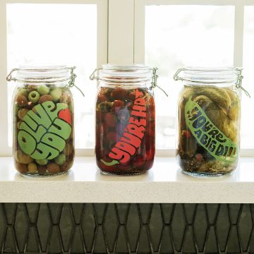 Show details for Brine & Dandy Three glass jars filled with olives, cherries, and pickles sit on a windowsill. Each jar has a playful label: "Oliv' You," "You're Cherry Sweet," and "You're a Big Dill.