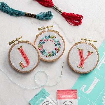 Show details for Holiday Letters Minis Embroidery Floss & Needle Three small embroidery hoops on a white surface, displaying the letters "J" and "Y" with a decorative floral wreath between them. Embroidery thread, a needle, and craft supplies are also visible.