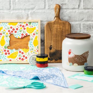 Show details for Mother Hen A farmhouse-style kitchen scene with chicken-themed items: a framed cutout, a wooden cutting board, a ceramic jar, paint, ink, scissors, a printed napkin, and a stencil, all set against a white brick wall.
