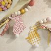 Two decorative bunny cutouts—one with a red floral pattern, the other with yellow plaid—each have a white pom-pom tail and are attached to a garland of pastel and gold beads, resting on a light stone surface.