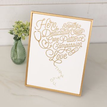 Show details for Healthcare Hero A gold-framed sign features words like “Hero,” “Dedication,” “Care,” “Patience,” “Compassion,” and “Love” arranged in a heart shape, along with medical-themed icons. A small vase with white flowers sits beside it.