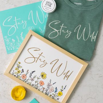 Show details for Stay Wild Encore Flat lay of a framed "Stay Wild" print with flowers, a matching green T-shirt, a turquoise clipboard, white ink, and yellow paint on a light surface. The script on each item reads "Stay Wild.