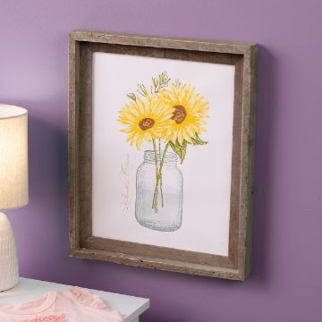 Show details for Mason Jar Sunflowers A framed picture of two yellow sunflowers in a mason jar hangs on a purple wall above a white table with a lamp and pink shirt.