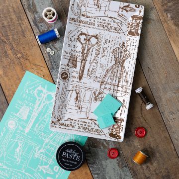 Show details for Vintage Sewing Pattern Flat lay of sewing-themed items on a wooden surface, including a patterned tray with dressmaking illustrations, sewing threads, buttons, a spool, a stencil sheet, blue chalk, and a jar of black paste.