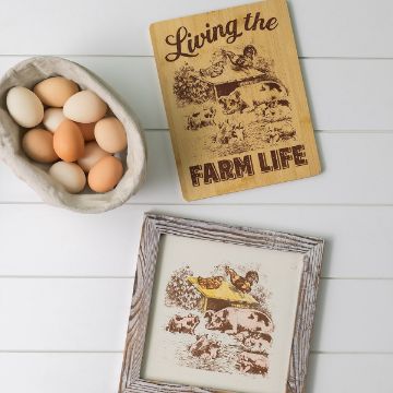 Show details for Living The Farm Life A bowl of brown and white eggs, a wooden plaque reading "Living the Farm Life" with farm animal illustrations, and a framed picture of similar farm animals on a white wooden surface.