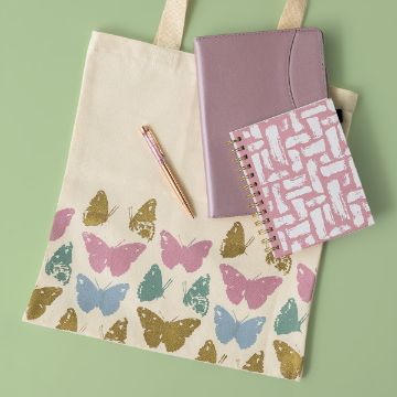 Show details for Butterfly Garden Patterns A beige tote bag with pink, blue, and gold butterfly prints, a pink and gold pen, a mauve planner, and a pink and white patterned spiral notebook are arranged on a light green background.