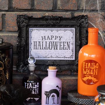 Show details for Gothic Happy Halloween A Halloween-themed display with a framed "Happy Halloween" sign, colorful potion bottles labeled "Halloween," and a pink bottle featuring a black cat and skeleton design, set against a brick wall.