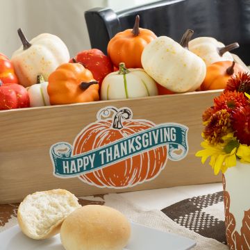 Show details for Thanksgiving Pumpkin A wooden box filled with decorative pumpkins sits on a table with a "Happy Thanksgiving" sign. In front, there are bread rolls and a cup with red and yellow flowers, creating a festive Thanksgiving setting.