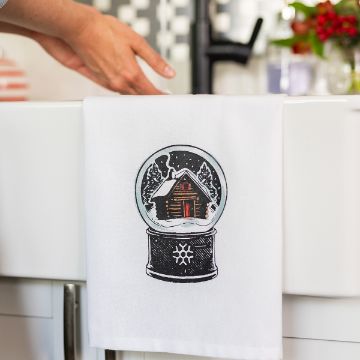 Show details for Log Cabin Snow Globe A white kitchen towel hanging over a sink features an illustration of a snow globe with a cozy cabin inside. A hand is reaching toward the sink in the background.