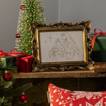 Show details for Holy Family A framed line drawing of the nativity scene sits on a wooden shelf, surrounded by wrapped gifts, a small decorated Christmas tree, ornaments, and a red pillow with white festive patterns.