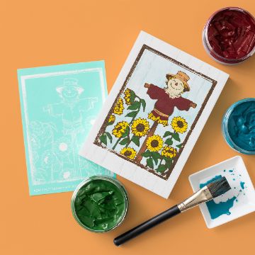 Show details for Scarecrow A painted scarecrow on a white wooden board surrounded by yellow sunflowers is displayed next to a stencil, jars of red, green, and blue paint, and a paintbrush on an orange background.