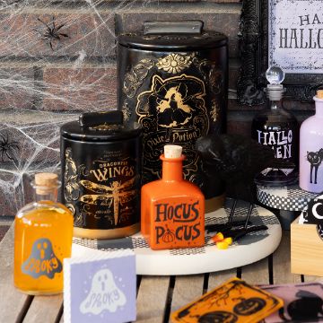 Show details for Twilight Apothecary A Halloween-themed display with potion bottles, black tins with gold labels, a crow figurine, faux cobwebs, and spider decorations. Bottles are labeled “Hocus Pocus,” “Spooky,” and “Dragonfly Wings.”.