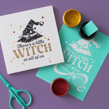 Show details for A Little Witch A square card with a witch hat illustration and the text “There’s a little WITCH in all of us” lies beside a teal art print of the same design, paint jars, a rubber stamp, and teal scissors on a purple surface.