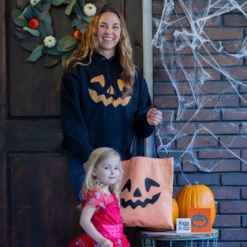 Show details for Happy Pumpkin A smiling woman in a black pumpkin face hoodie stands with a young girl in a red princess dress by a door decorated with fall leaves, fake cobwebs, pumpkins, and a "Hocus Pocus" sign, holding an orange pumpkin face tote bag.