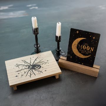 Show details for Moon Made Me Do It A small wooden table with a spiderweb and crescent moon design, two black candle holders with white candles, and a sign that reads "The moon made me do it" with a gold moon and stars.